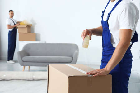 127055520 moving service employee sealing cardboard box with adhesive tape in room closeup